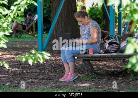 Kiew, Ukraine - 17. Juli 2026: Eine Frau arbeitet an einem Laptop und nutzt gleichzeitig ein Smartphone in einem schattigen Ruhebereich am VDNH (Expocenter) und genießt eine ruhige s Stockfoto
