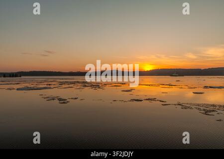 Ruhiger See bei Sonnenuntergang mit dünnem Eis, das auf stillem Wasser treibt, warmer orangefarbener Himmel und dunkle ferne Hügel schaffen eine friedliche Winterszenen in der Natur. Stockfoto