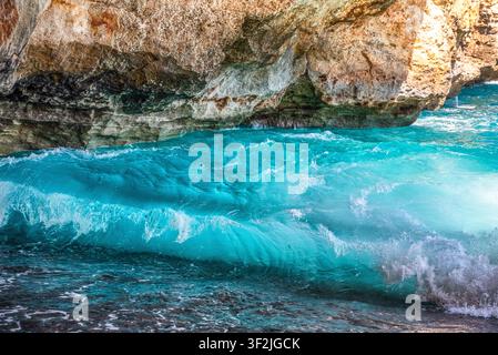 Nahaufnahme des kristallklaren türkisfarbenen Meeres mit sanften Wellen und einer felsigen Klippe im Hintergrund unter einem hellen Sommerhimmel Stockfoto