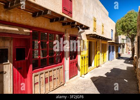 Sonnenbeschienene historische Straße auf der Insel Spinalonga, Kreta, mit restaurierten Steingebäuden, leuchtend roten Türen, gelben Fensterläden und traditionellem mediterranen Bogen Stockfoto