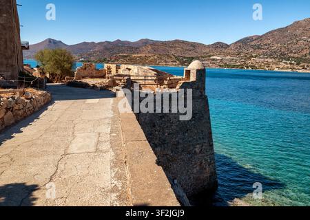 Historische venezianische Befestigungen und ein Küstenpromenade erheben sich über klarem türkisfarbenem Wasser auf der Insel Spinalonga, mit den zerklüfteten Bergen von Kreta im Stockfoto