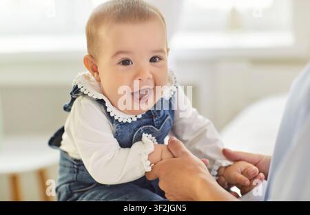 Zufriedenes Baby in Denim-Overall, das lächelt und zu Hause die Hände mit einem Elternteil hält Stockfoto