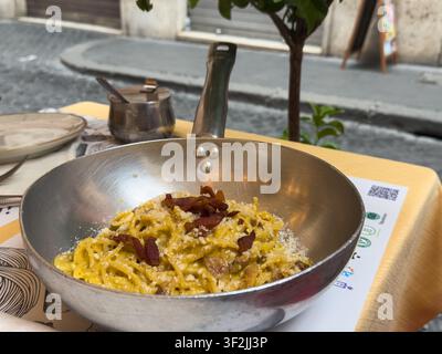 Authentische italienische Pasta Carbonara mit knusprigem Guanciale, serviert in einer traditionellen Metallpfanne auf einem Außentisch eines Restaurants in Rom, Italien. Stockfoto