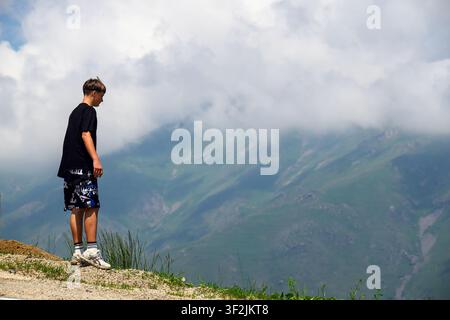 Junger Mann steht auf einem Bergrücken und blickt auf das grüne Tal. Wolken ziehen über entfernte Gipfel. Gras wächst nahe seinen Füßen. Die Szene zeigt eine natürliche Landschaft Stockfoto