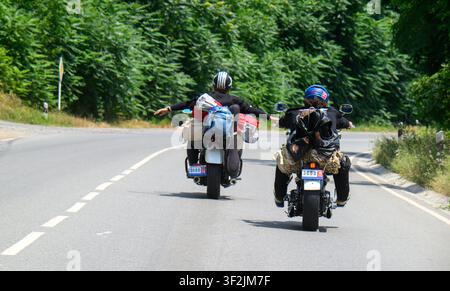 Zwei Motorradfahrer fahren auf einer kurvenreichen Straße durch ein Waldgebiet. Ein Fahrer gestikuliert mit einer offenen Hand. Der andere hält sich fest am Lenker Stockfoto