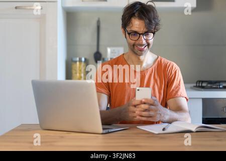 Zuhause, Mobiltelefon und Mann in der Küche, Laptop und Online-Lesen mit Kreativität, Projektforschung und Rezension. Soziale Medien, Freiberufler oder Person mit pc Stockfoto