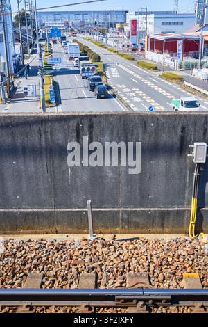 Blick auf eine erhöhte Eisenbahnschiene, der über eine belebte Vorstadtstraße und eine Betonbarriere blickt, perfekt für japanische Stadtinfrastruktur-Redaktionen Stockfoto