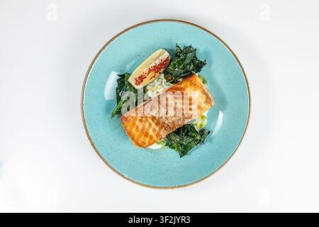 Gegrilltes Lachsfilet mit Spinat und Zitrone auf türkisfarbenem Teller, weißer Hintergrund, Top-Down-Ansicht. Stockfoto