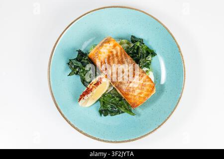 Eine Aufsichtsansicht eines gegrillten Lachsfilets mit Spinat, Erbsen und einer Zitronenscheibe auf einem gesprenkelten türkisfarbenen Keramikteller Stockfoto