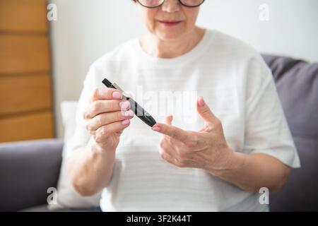 Zusammengeschnittene Aufnahme einer älteren Frau, die allein zu Hause ein Glukosemessgerät verwendet. Ungesunde, mittelalte, pensionierte Dame testet Blutzuckerspiegel am Morgen, sticht sich mit einer Lanzettenspritze in den Finger. Diabetes, Gesundheitskonzept Stockfoto