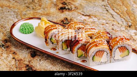 Ein farbenfrohes Teller mit Sushi-Rollen zeigt Schichten aus Fisch, Avocado und Frischkäse. Im stilvollen Restaurant serviert, ist das Gericht mit Wasabi und Ingwer in Scheiben garniert, was die Kunst des Kochens zur Schau stellt. Stockfoto