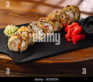 Goldgebratene Sushi-Rollen mit cremiger Füllung, Sauce und Sesamsamen liegen auf einer schwarzen Schiefertafel. Stockfoto