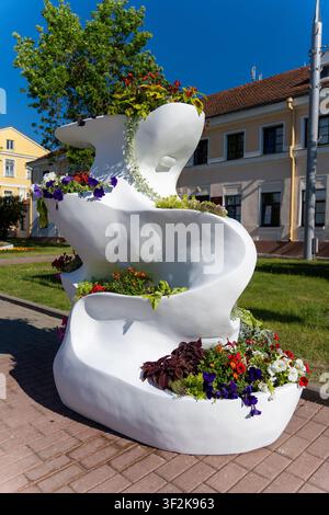 Farbenfrohe Blumen füllen skulpturale weiße urbane Blumenkästen entlang einer sonnigen Stadtstraße, mit historischer Architektur, geparkten Autos und einem blauen Verkehrsschild im Stockfoto