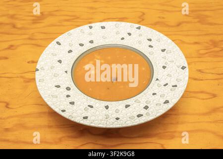 Dicke Orangensuppe mit sichtbaren Stücken, serviert in einer weißen Schale mit Mosaikrand auf einem Holztisch. Stockfoto