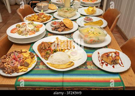 Vollständige Tischansicht eines peruanischen Festessens mit Lomo Saltado, Chicharrón, Suppen, Salaten und Dessert auf einem gestreiften Läufer in einem gemütlichen Restaurant. Stockfoto