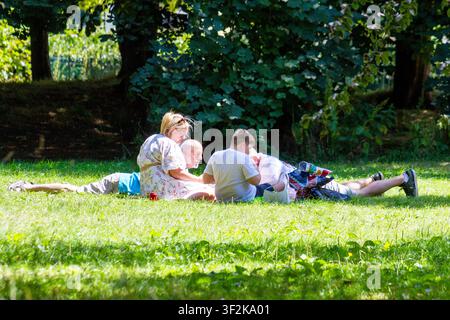 Eine Familiengruppe, die sich entspannt und miteinander spricht, während sie auf einem grünen Rasen in einem sonnigen Park sitzt und liegt. Southport, UK. Stockfoto