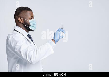 Afrikanischer Arzt in Gesichtsmaske und Handschuhen, die auf die Spritze in seiner Hand schauen, Seitenansicht, weißer Hintergrund, leerer Raum Stockfoto