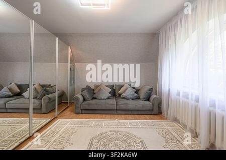 Gemütliches Zimmer mit einem grauen Sofa mit Kissen, einem gemusterten Teppich und einem großen Spiegel, der die Szene reflektiert. Natürliches Licht dringt durch den durchsichtigen weißen Vorhang Stockfoto