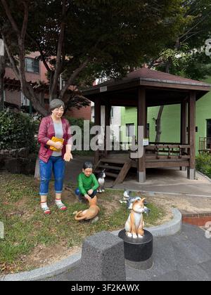 15. Juli 2025, Busan, Südkorea – Lebensgroße bemalte Figuren von Menschen und Hunden, die in einer ruhigen Wohnstraße ausgestellt sind, mit traditioneller Holztorstruktur. Stockfoto