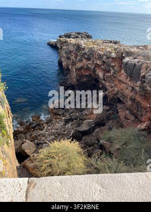 Luftartige Perspektive von einer Klippenwand aus, die über die schroffe felsige Mittelmeerküste mit tiefblauem Meer, Höhle und Küstenvegetation blickt, Europa. Stockfoto