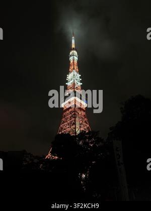 12. August 2025, Minato, Tokio, Japan – Tokyo-Turm leuchtet orange und weiß vor dunklem, wolkigem Nachthimmel, dramatische Froschaugensicht von unten aus gesehen. Stockfoto