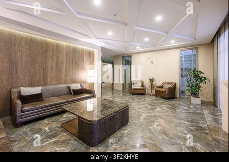 Elegante Lobby mit einem braunen Sofa, Sesseln, Marmorböden und einem einzigartigen Deckendesign. Warme Beleuchtung unterstreicht die raffinierte Atmosphäre Stockfoto