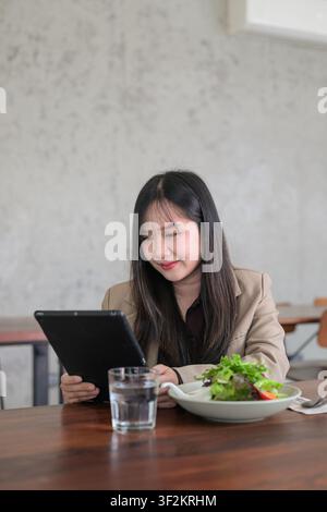 Ein vertikales Bild zeigt eine junge asiatische Frau, die mit einem konzentrierten und dennoch sanften Lächeln auf ein schwarzes Digitaltablett schaut. Gekleidet in einem professionellen Beige Stockfoto