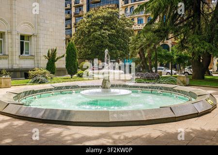 Batumi, Georgien – 09. Juni 2026: Antike Steinfontäne mit kreisförmigem Becken und Wasserstrahlen in einem öffentlichen Park, umgeben von üppigen tropischen Bäumen Stockfoto