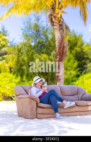 Reisender entspannt sich auf einem Sofa, das auf einem tropischen Strand unter Palmen platziert ist, während er ein Smartphone auf Maamigili Island, Malediven, nutzt. Stockfoto