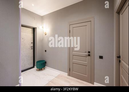 moderner Flur mit grauen Wänden, zwei beigen Türen, gefliestem Boden, grünem Hocker und einer stilvollen Wandleuchte Stockfoto