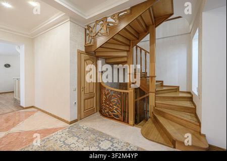 Elegantes Foyer zeigt eine hölzerne Wendeltreppe mit kunstvollen Schnitzereien. Ein gemusterter Teppich liegt auf dem Fliesenboden, mit einer Holztür in der Nähe Stockfoto