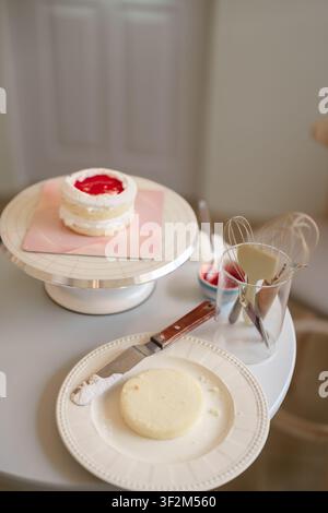 Hausgemachter geschichteter Kuchen auf elegantem Kuchenständer mit Rührutensilien und Zutaten, die auf weißer Thekenoberfläche ausgebreitet sind Stockfoto