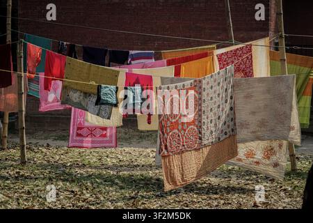 Verschiedene farbenfrohe Stoffe, darunter rot, orange, rosa, grün und gemusterte Textilien, hängen an einer Wäscheleine, die zwischen zwei Bambusstäben gespannt ist. Der Boden Stockfoto