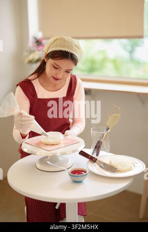 Asiatische Frau streicht Creme auf einen Kuchen an einem weißen Tisch in einer hellen Küche mit Fensterlicht und Backutensilien Stockfoto