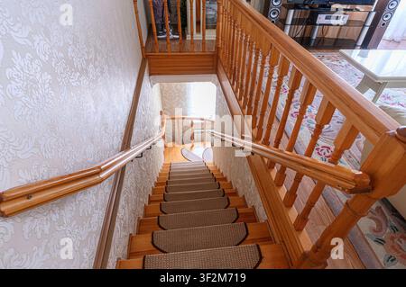 Hohe Winkelaufnahme, die eine Holztreppe mit Teppichstufen hinunterblickt. Die Wände sind mit einem Blumenmuster tapeziert. Holzgeländer befinden sich auf beiden Seiten von Stockfoto