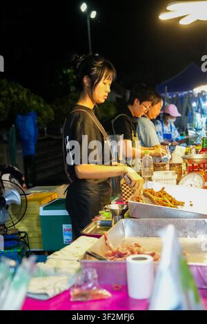 Nan Thailand: Junge Straßenhändlerin, die frittiertes Essen mit einer Zange an einem geschäftigen Nachtmarktstand serviert Stockfoto