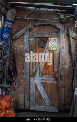 Alte hölzerne Brettertür mit diagonalen Streben, blauem Seil und „I LOVE YOU“-Aufkleber, eingebettet in einer OSB-Wand-Werkstatt mit Schläuchen, Seilen und Stöcken Stockfoto