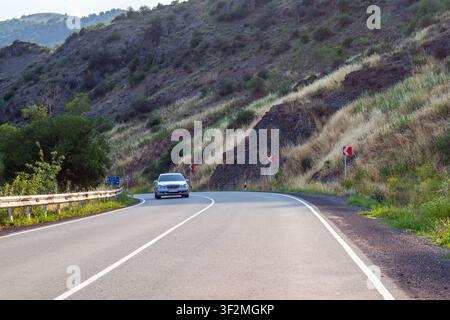 Weißes Auto fährt auf einer kurvenreichen Bergstraße. Die Straße windet sich durch trockene, grasbewachsene Hänge. Ein Geländer säumt die linke Straßenseite Stockfoto