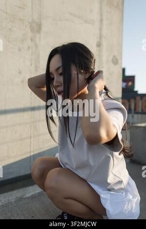 Nahaufnahme einer stilvollen asiatischen Frau, die auf einem städtischen Dach sitzt und ihr langes Haar im warmen Goldlicht der Abendstunde richtet, mit einem Betonhintergrund. Perfekt für Stockfoto