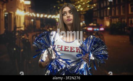 Eine junge Cheerleaderin, die blaue Metallic-Pompons hält, die Hände sichtbar, auf einer von Lichtern gesäumten Straße schmeichelt; Langeweile Einsamkeit. Stockfoto