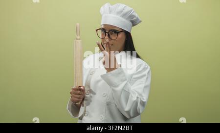 Junge hispanische Köchin hält einen hölzernen Nudelholz und zeigt zwei Finger zu ihrem Gesicht im Studio; selbstbewusstes Handwerk. Stockfoto