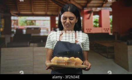 Bäckerin, die ein Tablett mit Muffins hält und grimmig macht, während sie sie in einem Gebäude präsentiert; ekelhafte Authentizitätsarbeit. Stockfoto