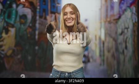 Frau, die in einer Graffiti-Straßenallee Daumen mit sichtbaren Daumen hochlegt und ein breites Lächeln trägt, in Creme-Pullover und Jeans, mit Blick auf die Kamera; Glück Stockfoto