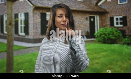 Frau Brünette mittleren Alters in gestreiftem Hemd zeigt den Zeigefinger auf die Kamera vor einem Ziegelgebäude mit Rasen und Fensterläden; durchsetzungsstark. Stockfoto
