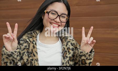 Frau blitzt Friedenszeichen mit beiden Händen, trägt Leopardenfellmantel an einer hölzernen Gebäudewand; verspielte Freude. Stockfoto