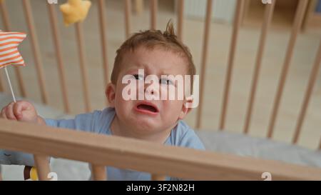 Kleinkind Junge hispanic weint, greift nach orange gestreiftem Sternenspielzeug und packende Gitterstange in hölzernem Kinderbett im Gebäude; Distress braucht Komfort. Stockfoto