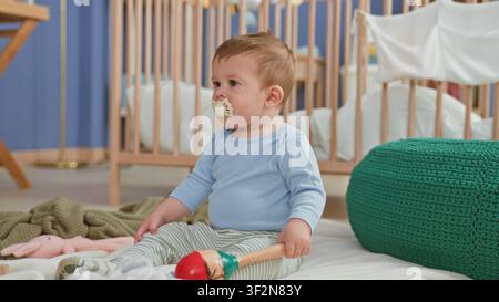 Kleiner Junge sitzt mit Schnuller und hält eine Maracas, streckt die Hand in Richtung Bett in einem Gebäude; unschuldige Neugier. Stockfoto