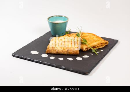 Studio Hochwinkelaufnahme von goldenen gefalteten Pfannkuchen, serviert mit Sauerrahm in einer türkisfarbenen Tasse, frische Mikrogrüns Stockfoto
