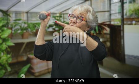 Seniorin, die eine 1-Kilogramm-Kurzhantel hält und mit dem Finger auf sie in einem Gewächshaus zeigt; Fitness Vitalität Selbstvertrauen. Stockfoto