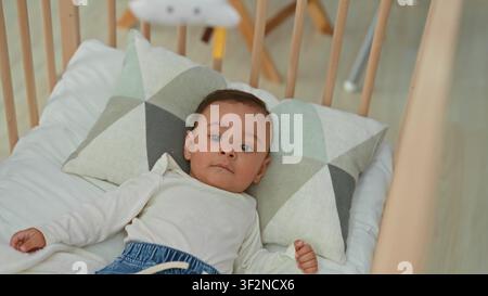 Hispanisches Baby, das in einer Holzwiege auf einem geometrischen Kissen liegt, Hände sichtbar und Kopf gedreht, schaut nach oben in einem Gebäude; ruhige Neugier. Stockfoto